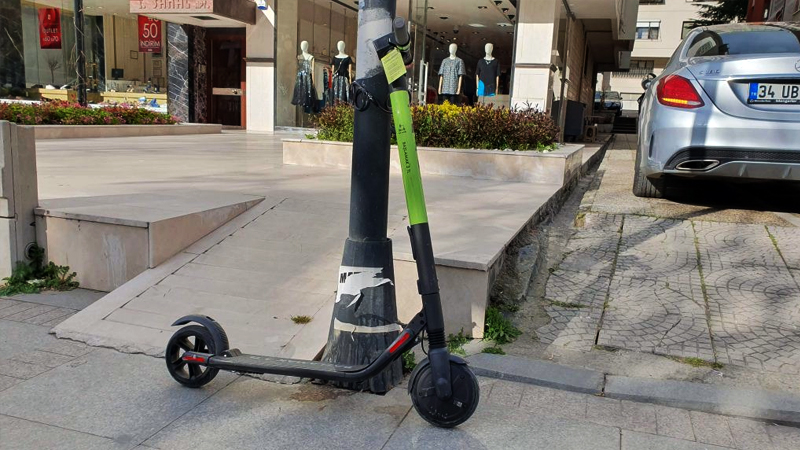 İstanbul'da kaldırımda bir direğe bağlanmış elektrikli scooter. Arka planda bir mağazanın vitrini ve vitrinin sağında bir araba görünüyor.
